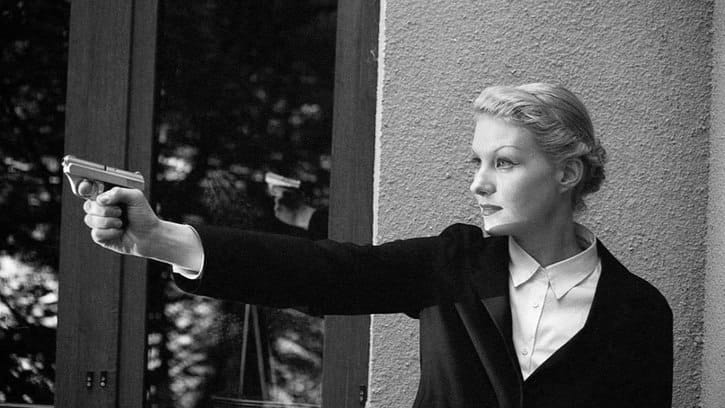 Black-and-white photograph of a woman with pinned-up light hair, wearing a dark jacket and white shirt, aiming a pistol.
