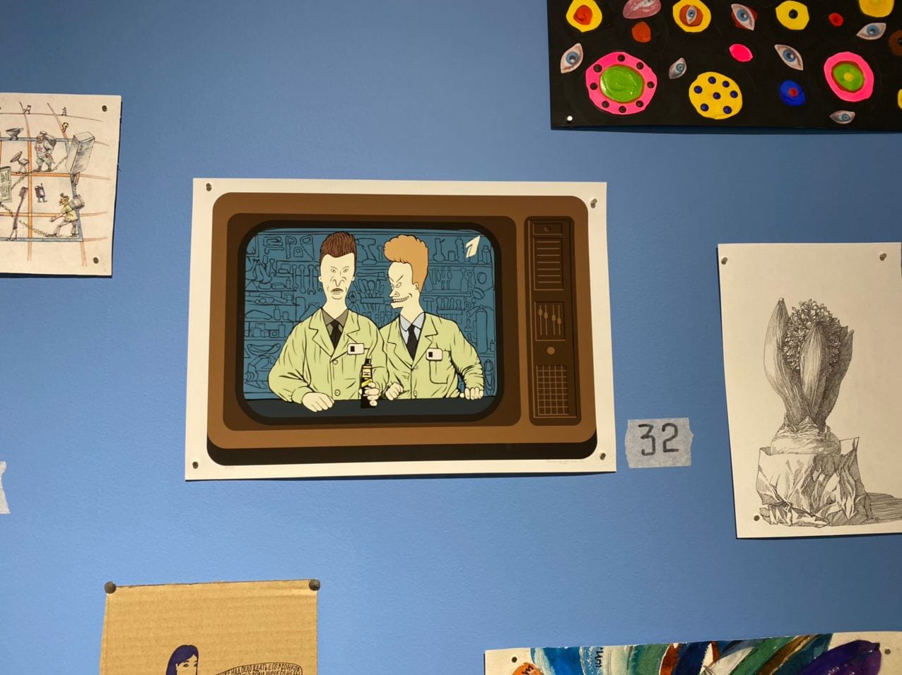 Sur un mur bleu, une estampe montre deux personnages de dessin animé en blouse claire et cravate sur un téléviseur rétro. Une étiquette indique 32 ; d'autres dessins sont accrochés autour.