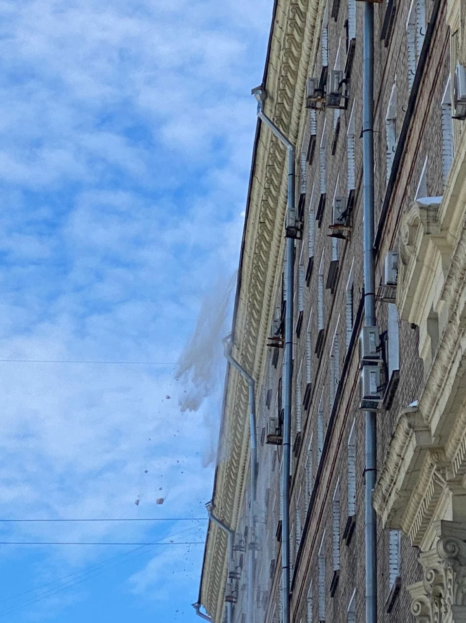 Vue en contre-plongée d'une façade avec gouttières et climatiseurs. De la neige et des morceaux de glace tombent du toit sous un ciel nuageux.