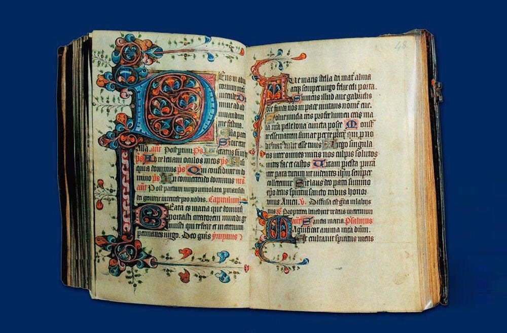 An open medieval book of hours with Latin text and ornamented blue and red initials, set against a dark blue background.