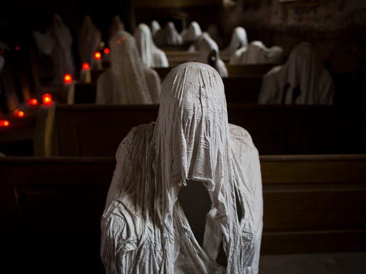 Une figure blanche au visage couvert de tissu est assise sur un banc en bois dans une église sombre. Derrière elle, des rangées de figures semblables et des bougies rouges.