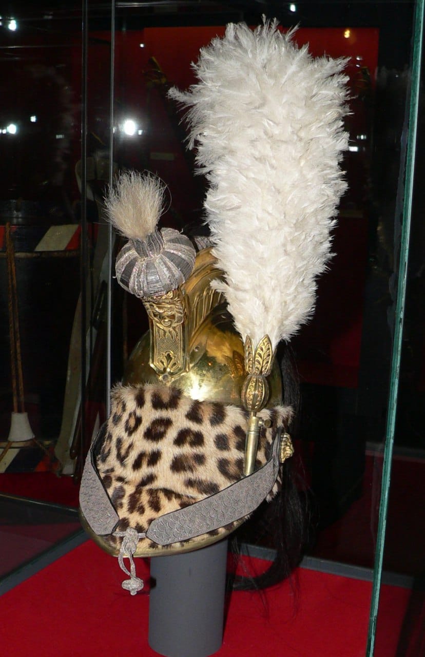 Casque en laiton bordé de peau de léopard, avec un haut plumet blanc et un pompon argenté, sur un socle derrière une vitre, sur fond rouge.