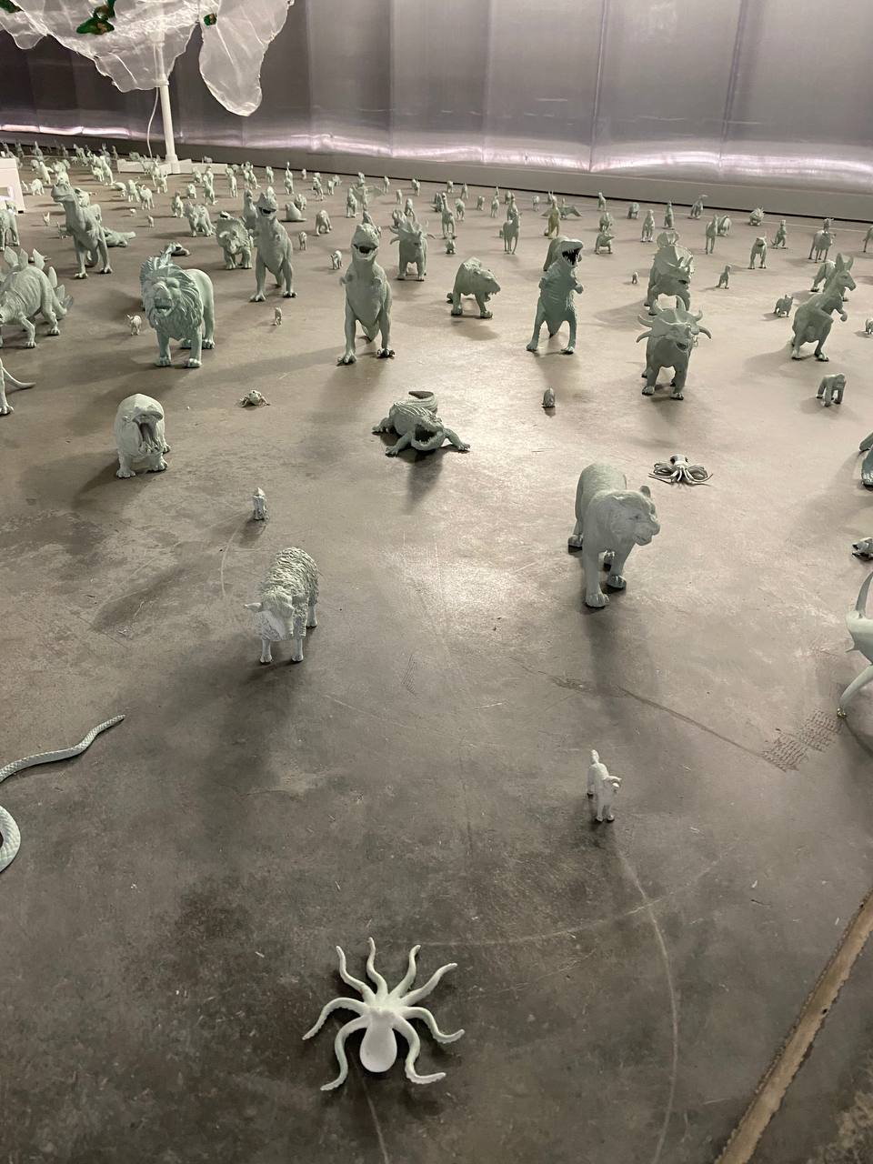 Des centaines de figurines monochromes sur un sol en béton : lions, dinosaures, crocodile, mouton, kangourou, serpent. Au premier plan, une pieuvre blanche ; un tissu translucide en haut.