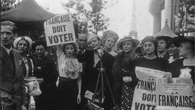 Photo en noir et blanc de femmes en chapeaux avec une pancarte « Française doit voter » et des journaux La Femme nouvelle, la tour Eiffel en arrière-plan.