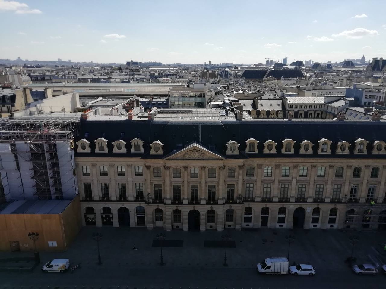 Façade en pierre à fronton triangulaire et lucarnes sous un toit sombre. Échafaudages à gauche, voitures sur la place en bas, panorama de la ville sous un ciel clair.