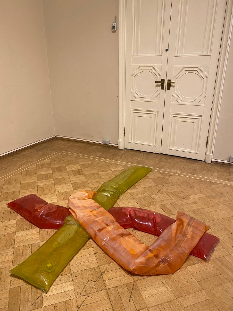 Des poches translucides rouges, jaune-vert et orange s'entrecroisent sur un parquet. Au fond, une porte blanche à deux battants aux panneaux moulurés.