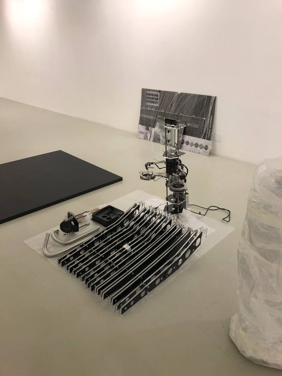 Dans une salle blanche de galerie, des plaques noires perforées reposent sur du papier bulle près d'un mécanisme métallique dressé, avec des fils. Un dessin noir et blanc est appuyé au mur.
