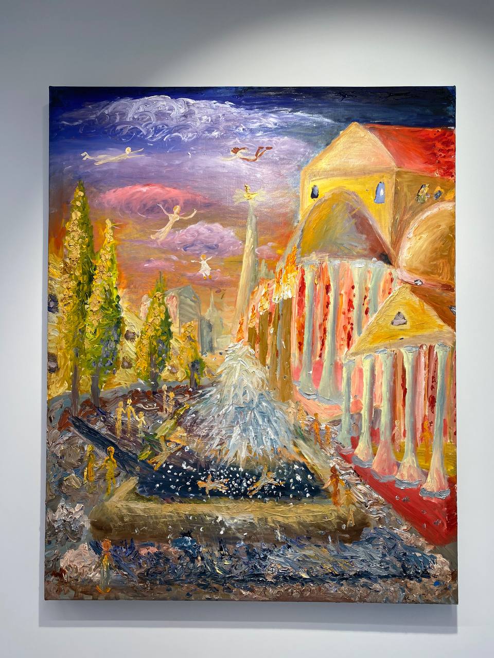 Tableau accroché au mur, en touches épaisses : bâtiments jaunes et rouges à colonnes, fontaine blanche, cyprès, petites figures en bas et silhouettes volant dans un ciel lilas.