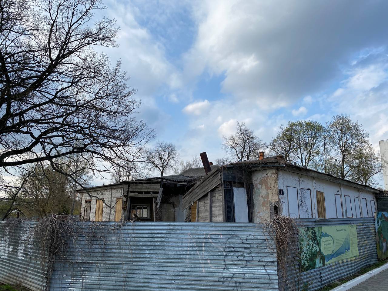 Maison délabrée de plain-pied aux fenêtres condamnées et au toit affaissé, derrière une clôture en tôle ondulée. Arbres nus et ciel nuageux.
