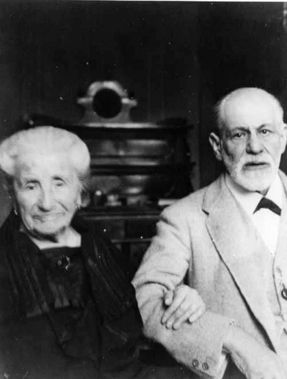 Photographie en noir et blanc d'une femme âgée en vêtements sombres et d'un homme à barbe grise en veste claire, assis côte à côte.