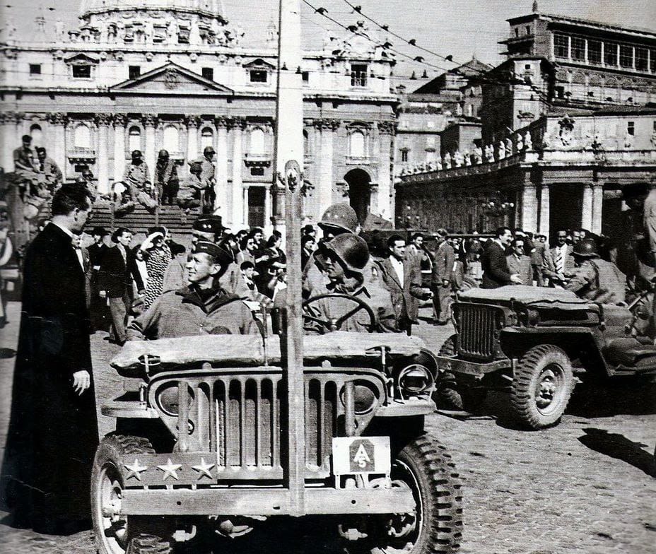 Des jeeps militaires avec des soldats américains casqués devant la basilique Saint-Pierre. À gauche, un prêtre en soutane ; une foule de civils les entoure.