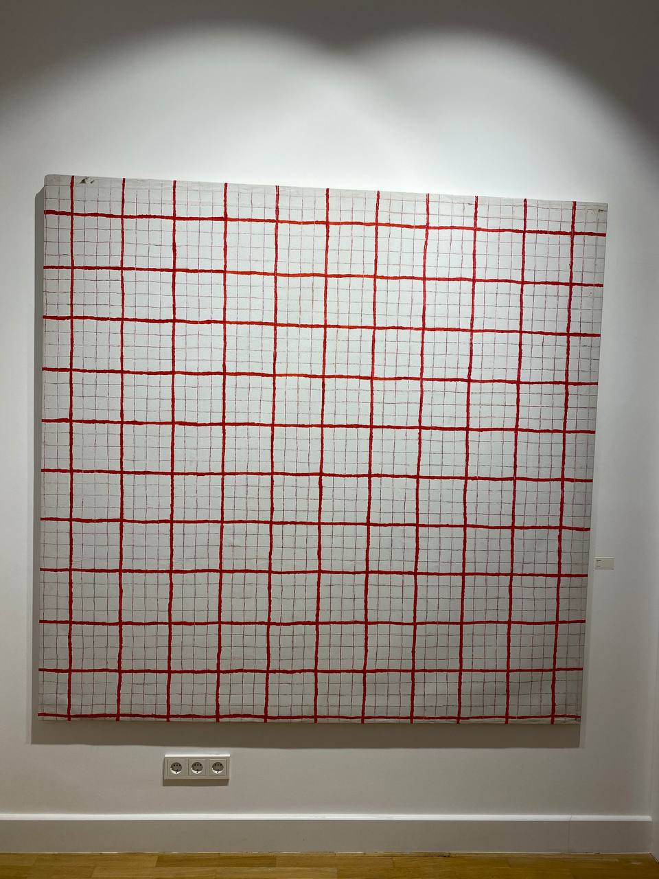Toile carrée sur un mur blanc : fond blanc et quadrillage de lignes rouges, épaisses en alternance avec des fines. Trois prises en dessous, un petit cartel à droite.
