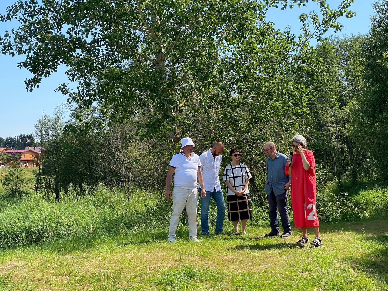 Cinq personnes se tiennent sur l'herbe sous un grand arbre, par temps ensoleillé. Une femme en robe rouge parle au micro, les autres écoutent. Une maison en bois au loin.