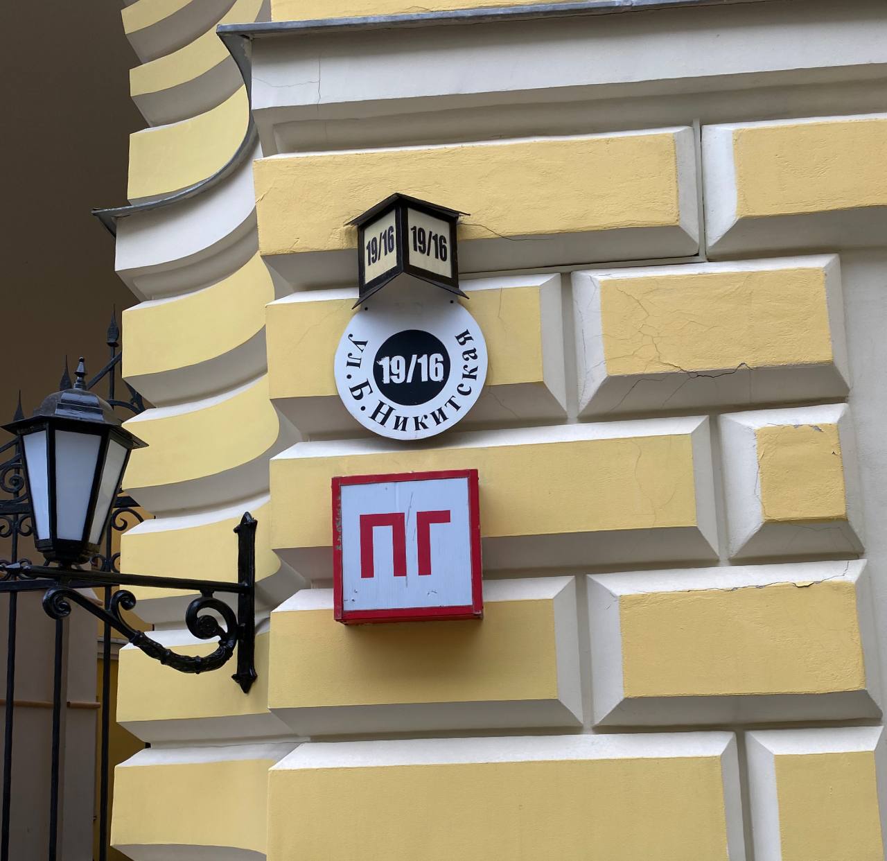 Corner of a yellow rusticated building with a round plaque reading Bolshaya Nikitskaya Street 19/16, a lantern showing the number and a red and white PG sign.