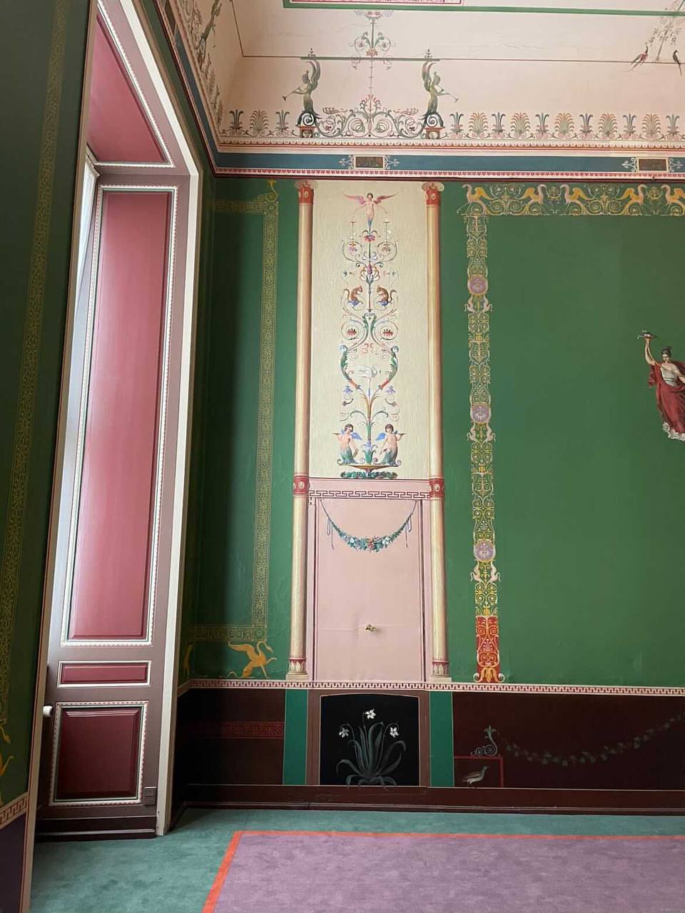Angle d'un intérieur aux murs verts peints d'arabesques, de putti et de griffons. Panneau orné entre deux fines colonnes, figure en rouge à droite, porte rose à gauche.