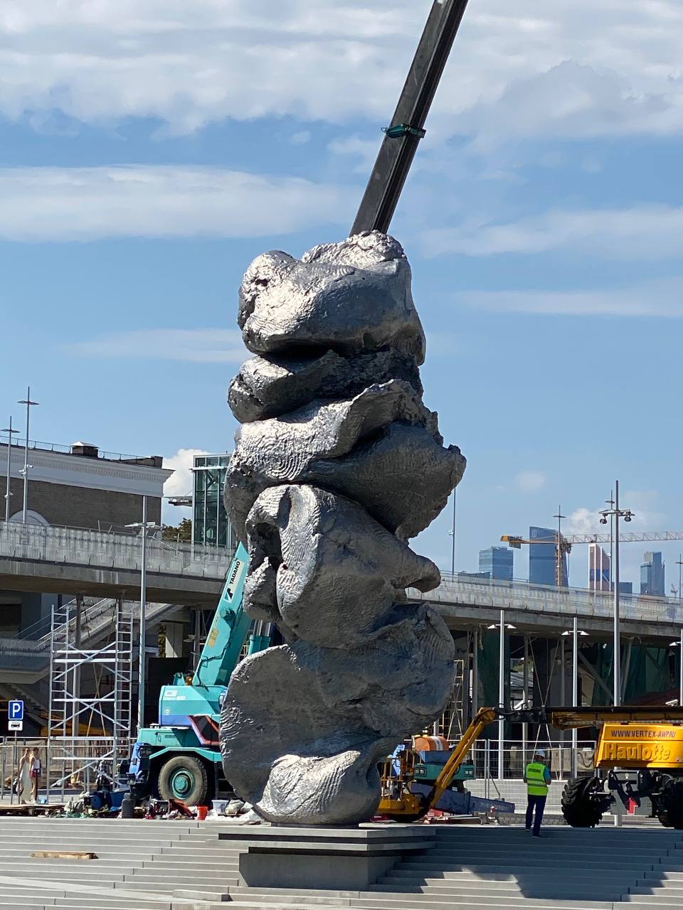 A twelve-metre aluminium sculpture of stacked, irregular lumps on a square, with a mobile crane and a lift beside it.