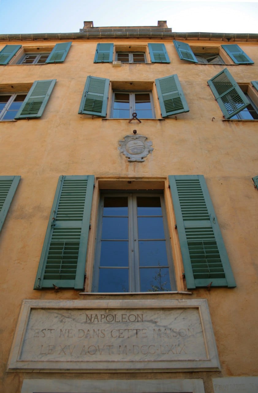 Ochre house facade with green shutters and a marble plaque recording Napoleon's birth on 15 August 1769, seen from below.