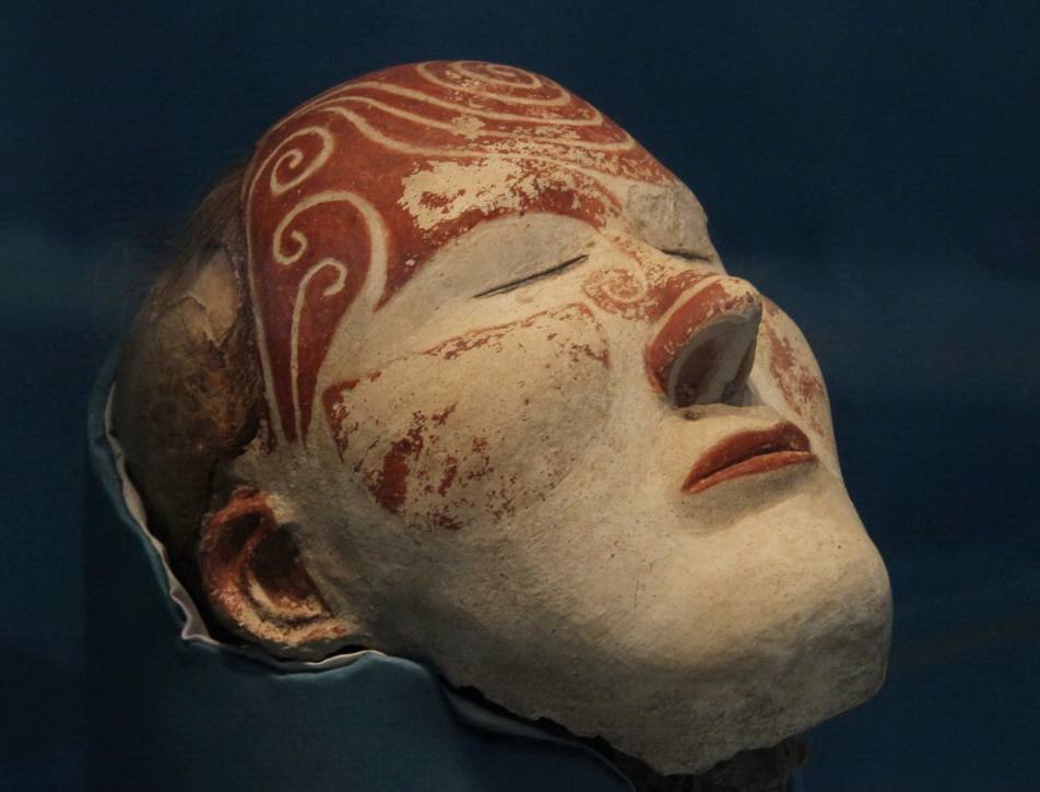 Tête aux yeux clos recouverte d'enduit blanc, ornée de spirales rouges sur le front et les joues, en partie effacées. Fond bleu sombre.