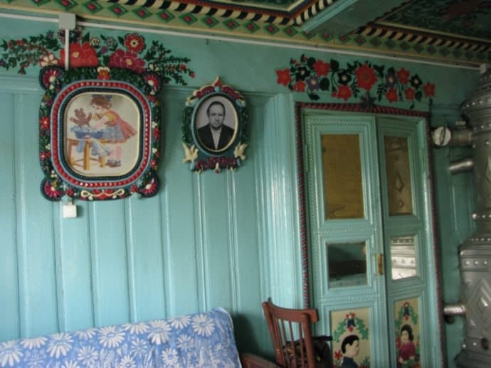 Pièce aux murs turquoise ornés de fleurs rouges. Deux portraits dans des cadres ornés près d'une porte peinte, un poêle métallique à droite, une chaise et un canapé en bas.