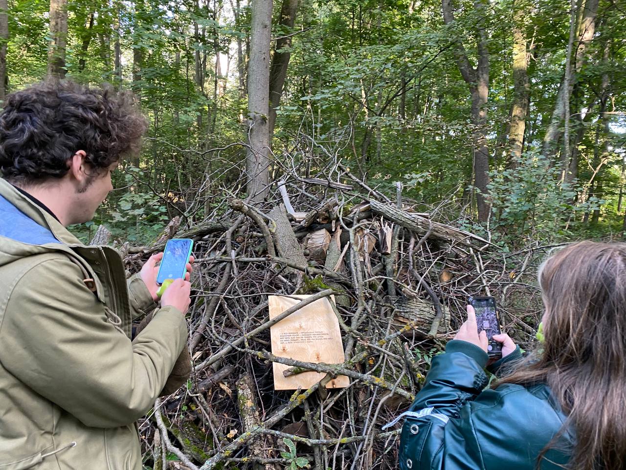 Un jeune homme et une jeune femme photographient avec leur smartphone une plaque de contreplaqué portant du texte, fixée sur un grand tas de branches dans une forêt estivale.