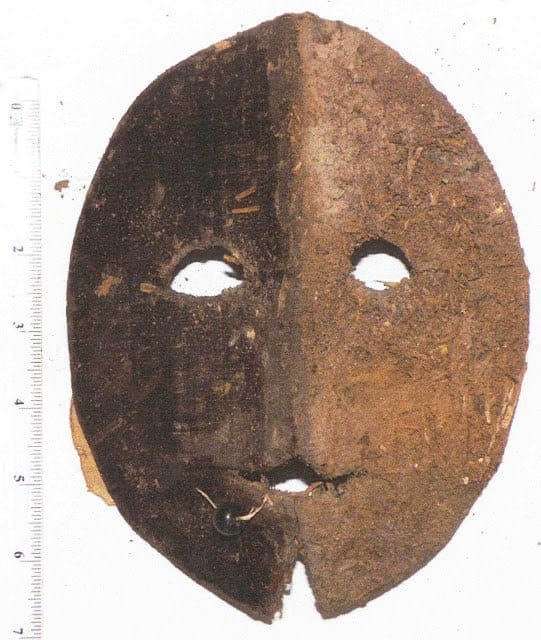 Masque ovale percé pour les yeux et la bouche, moitié gauche plus sombre que la droite, perle près de la bouche, règle à gauche.
