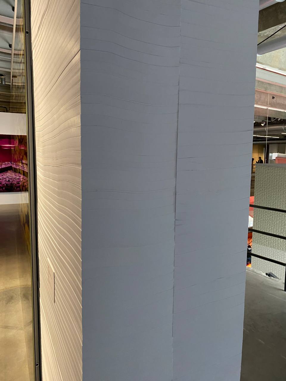 Colonne gris-bleu faite de fines couches horizontales, comme une pile de papier. À gauche, derrière la vitre, une salle montre une œuvre rose et violette sur le mur.