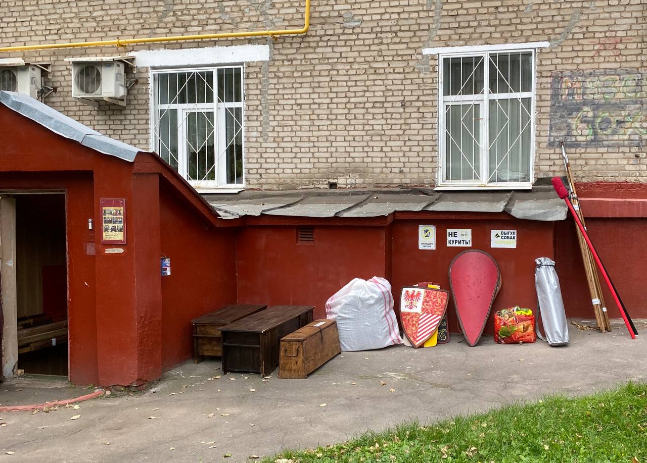 Chests, knightly shields, bags and poles beside a red basement entrance of a brick building.