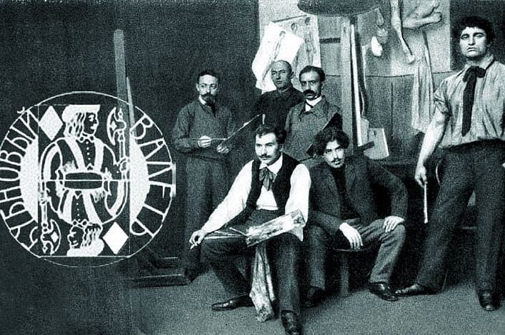Photographie de groupe en noir et blanc de sept hommes dans un atelier, avec à gauche un emblème rond représentant un valet de carte et l'inscription « Bubnovy valet ».
