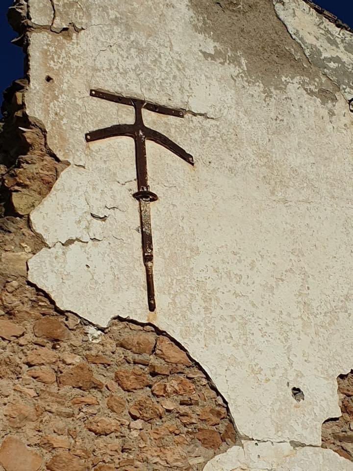 A dark iron anchor shaped like a cross on a stone wall with peeling white plaster.