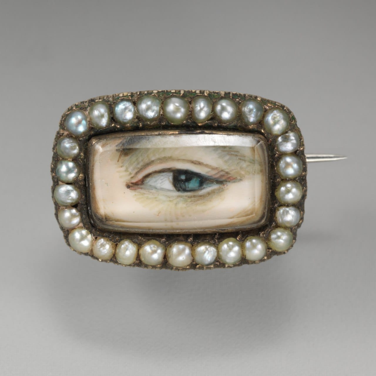 Rectangular brooch with a painted eye under glass, surrounded by a row of small pearls, with the pin visible on the right.