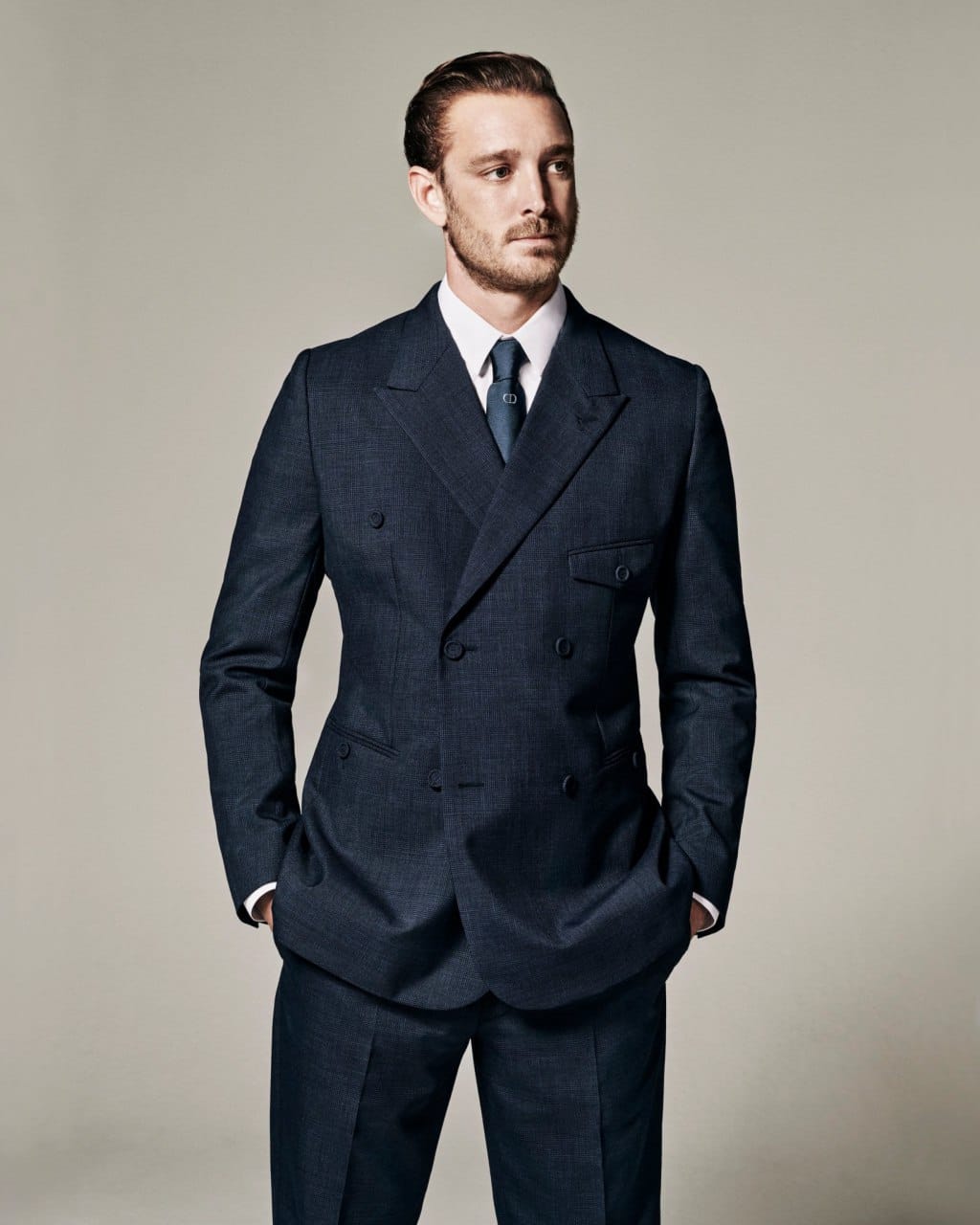 Un jeune homme à la barbe courte, en costume croisé bleu marine, chemise blanche et cravate bleue, se tient de trois quarts, les mains dans les poches. Fond gris-beige.