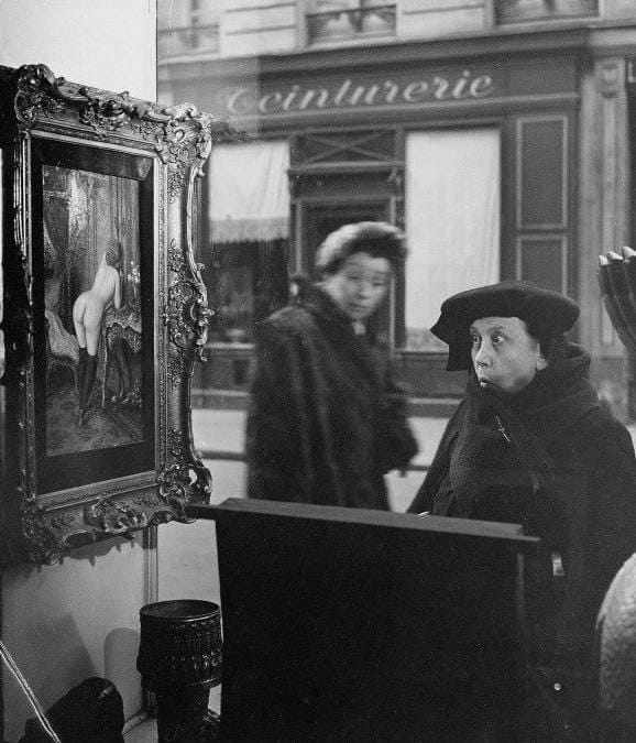 Black-and-white photograph of an older woman in a dark hat and coat looking at a painting of a nude in a shop window, with the street reflected in the glass.