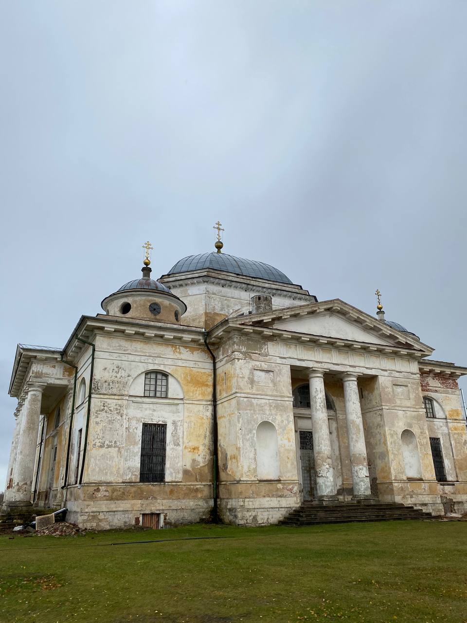 Église de style classique avec colonnes de portique, dôme gris et croix dorées. Façade au crépi écaillé, pelouse au premier plan.
