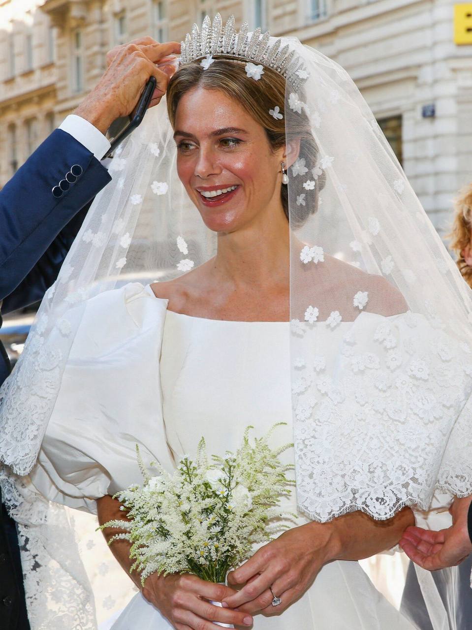 Une mariée souriante en robe blanche à manches bouffantes, diadème et long voile fleuri tient un bouquet ; une main ajuste le voile. Un immeuble en arrière-plan.