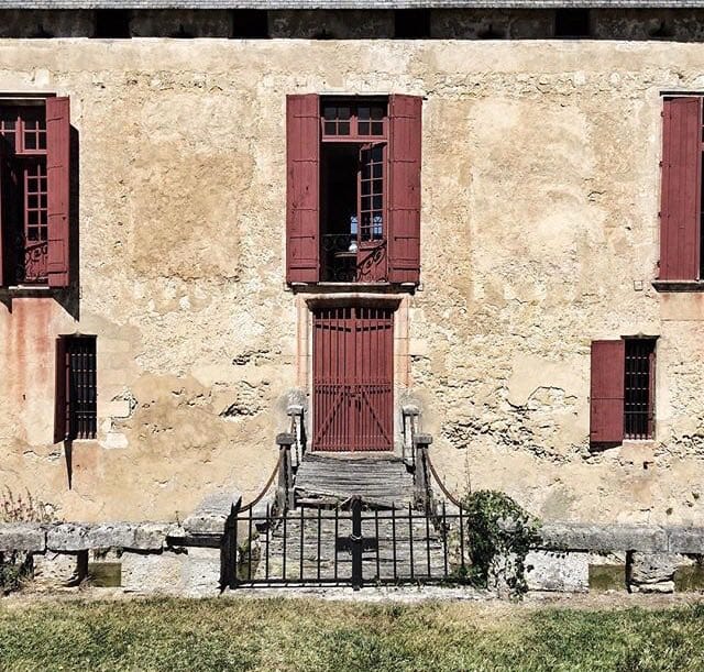 Facade of an old castle in yellowish stone with red shutters, a red door and a small bridge with an iron gate in front of the entrance.