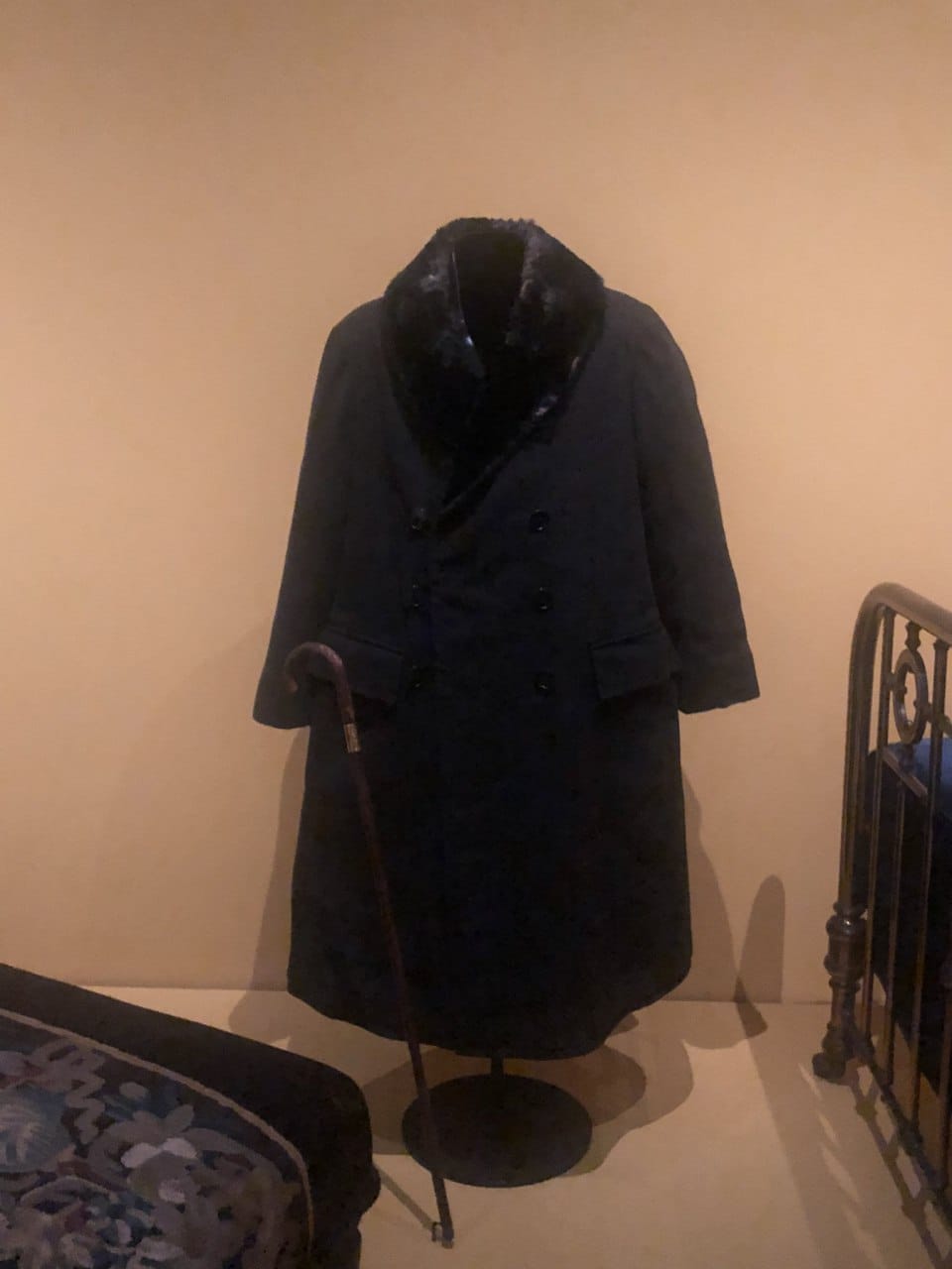 A dark double-breasted coat with a fur collar on a stand, a cane beside it and a metal bed frame at right, in a museum interior.