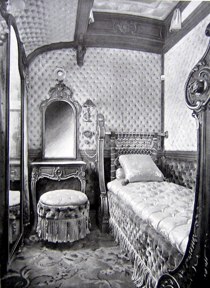 Photo en noir et blanc d'un petit compartiment : murs capitonnés, lit à franges avec coussin, pouf, coiffeuse à miroir, tapis à motifs, miroir à gauche.