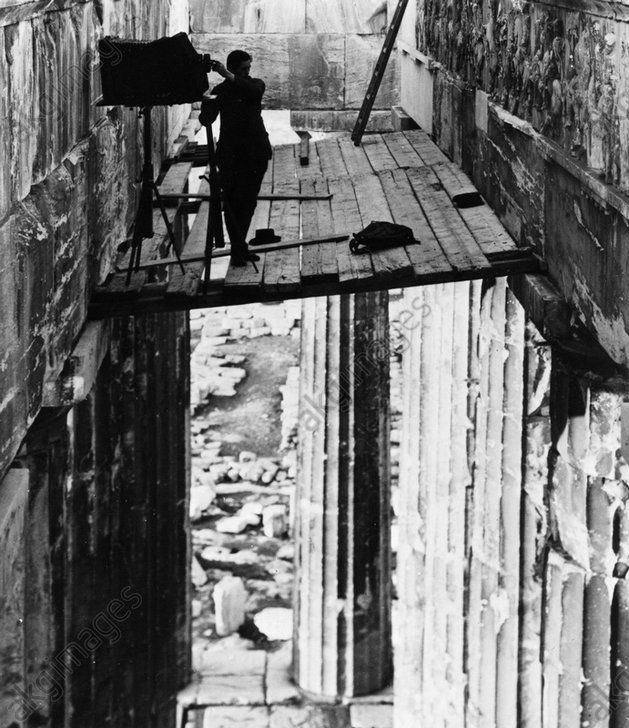Photo en noir et blanc : un photographe se tient près d'un appareil sur trépied, sur une plateforme en bois entre des murs de pierre, au-dessus de colonnes cannelées.