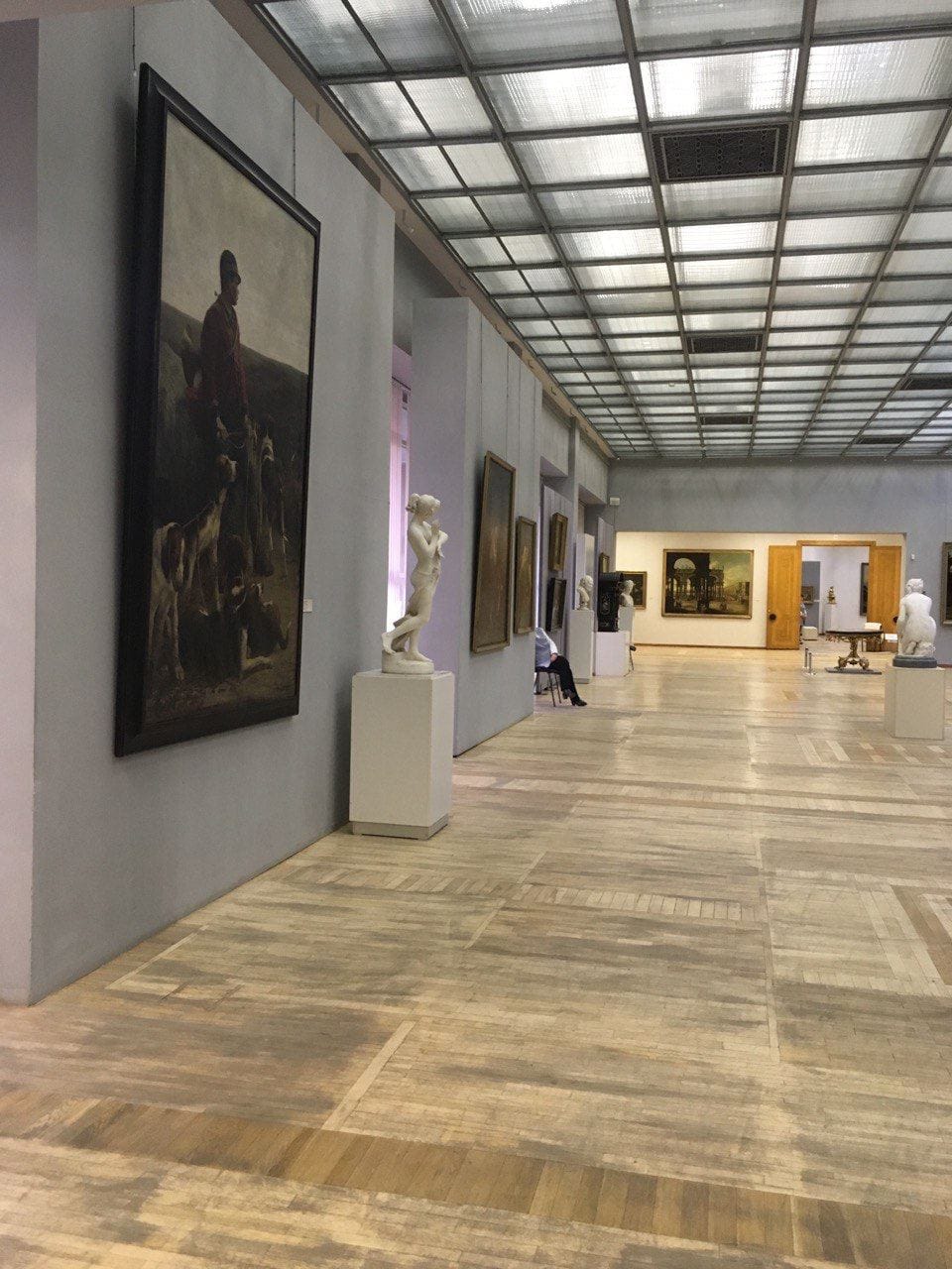 Vaste salle de musée au parquet et au plafond vitré. À gauche, un grand tableau d'un chasseur avec ses chiens, près d'une statue de marbre sur un socle.