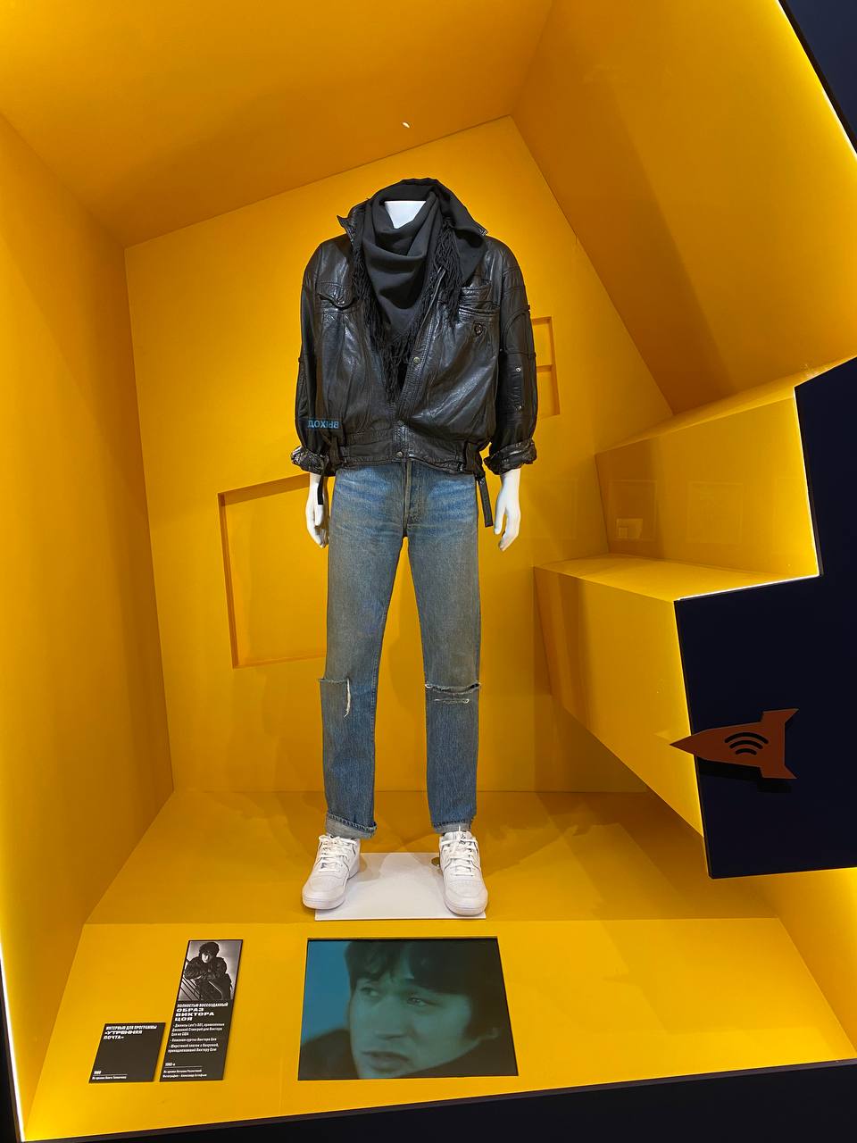 Un mannequin en blouson de cuir noir, foulard sombre, jean usé et baskets blanches dans une vitrine jaune. En bas, une étiquette et un écran diffusant une interview.