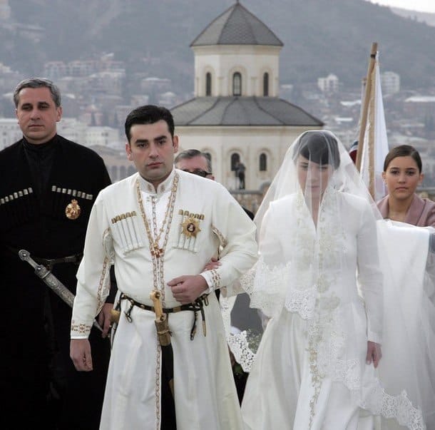 A groom in white traditional Georgian dress with a dagger and a bride in a white gown with a long veil walk side by side before a hilltop church.