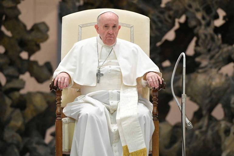 An elderly man in white papal vestments with a pectoral cross sits in an armchair, with a microphone beside him.