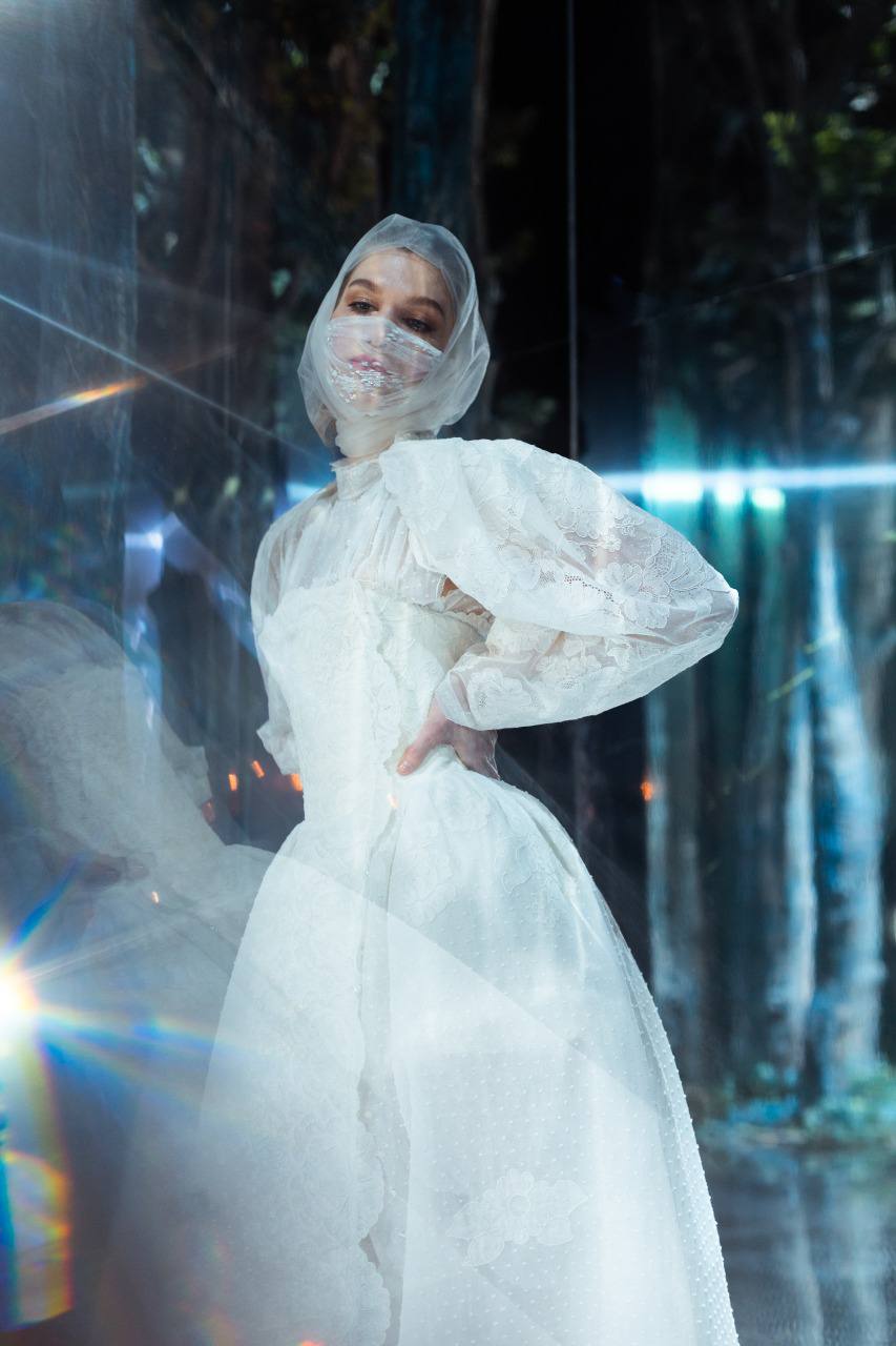 Une jeune femme en robe de dentelle blanche à manches bouffantes, capuche transparente et masque pose la main sur la hanche. Reflets bleutés, éclats de lumière et fond de forêt sombre.