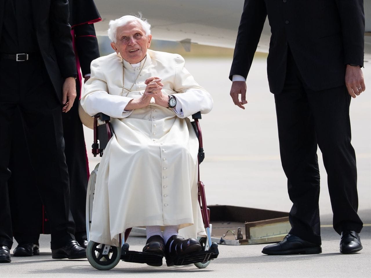 Un vieil homme aux cheveux blancs, en soutane blanche, est assis dans un fauteuil roulant, les mains jointes, le regard levé. Des hommes en costume sombre l'entourent, un avion derrière.