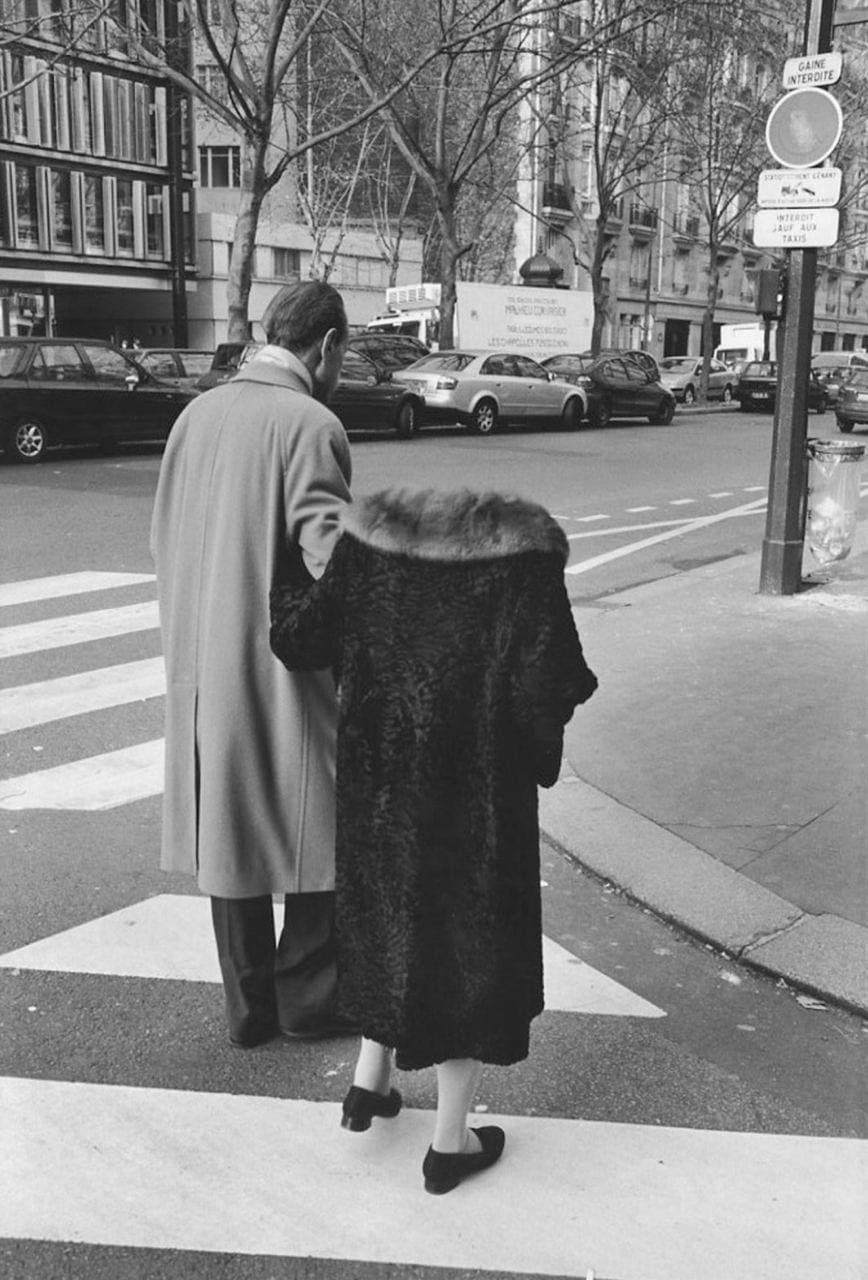 Black-and-white photograph of an elderly man in a coat and a woman in a fur coat, seen from behind, crossing a Paris street at a pedestrian crossing.