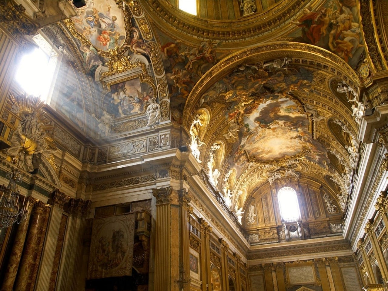 Intérieur d'église baroque : voûte ornée de fresques, de stucs dorés et de sculptures blanches, avec des rayons de soleil par la fenêtre à gauche.