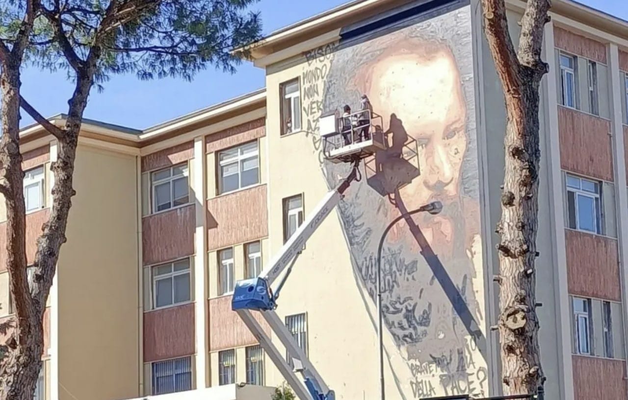 Un immense portrait d'homme barbu, accompagné d'inscriptions en italien, orne le mur d'un immeuble. Deux ouvriers se tiennent sur une nacelle élévatrice, entre des troncs de pins.