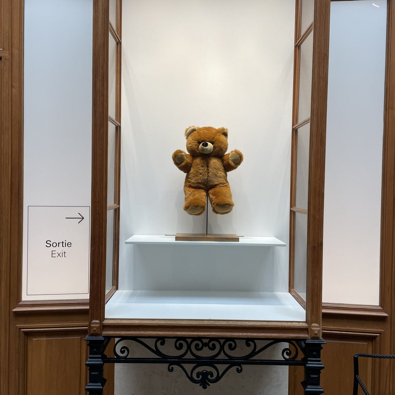 A teddy bear on a stand inside a wooden display case with white walls, with a Sortie Exit sign and arrow on the left.