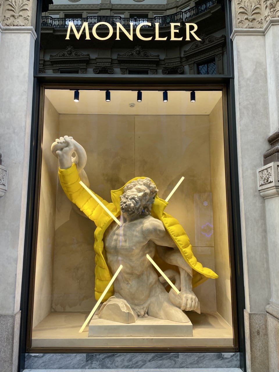 Short Aspin down jacket in buttercup yellow and antique sculptures in the windows of the Moncler boutique in the Galleria Vittorio Emanuele II, Milan