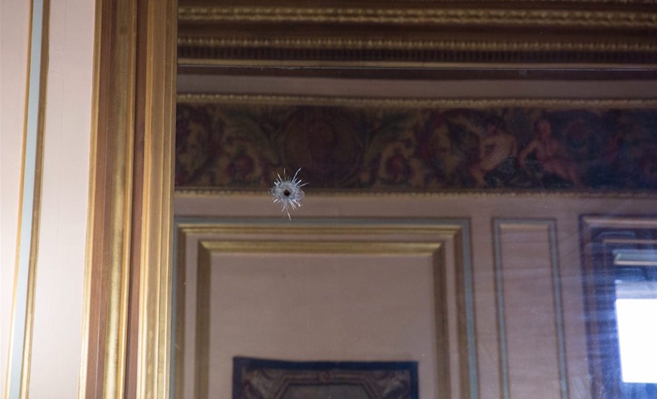 A mirror in a gilded frame with a bullet hole and cracks, reflecting the moulded and painted interior of a palace.